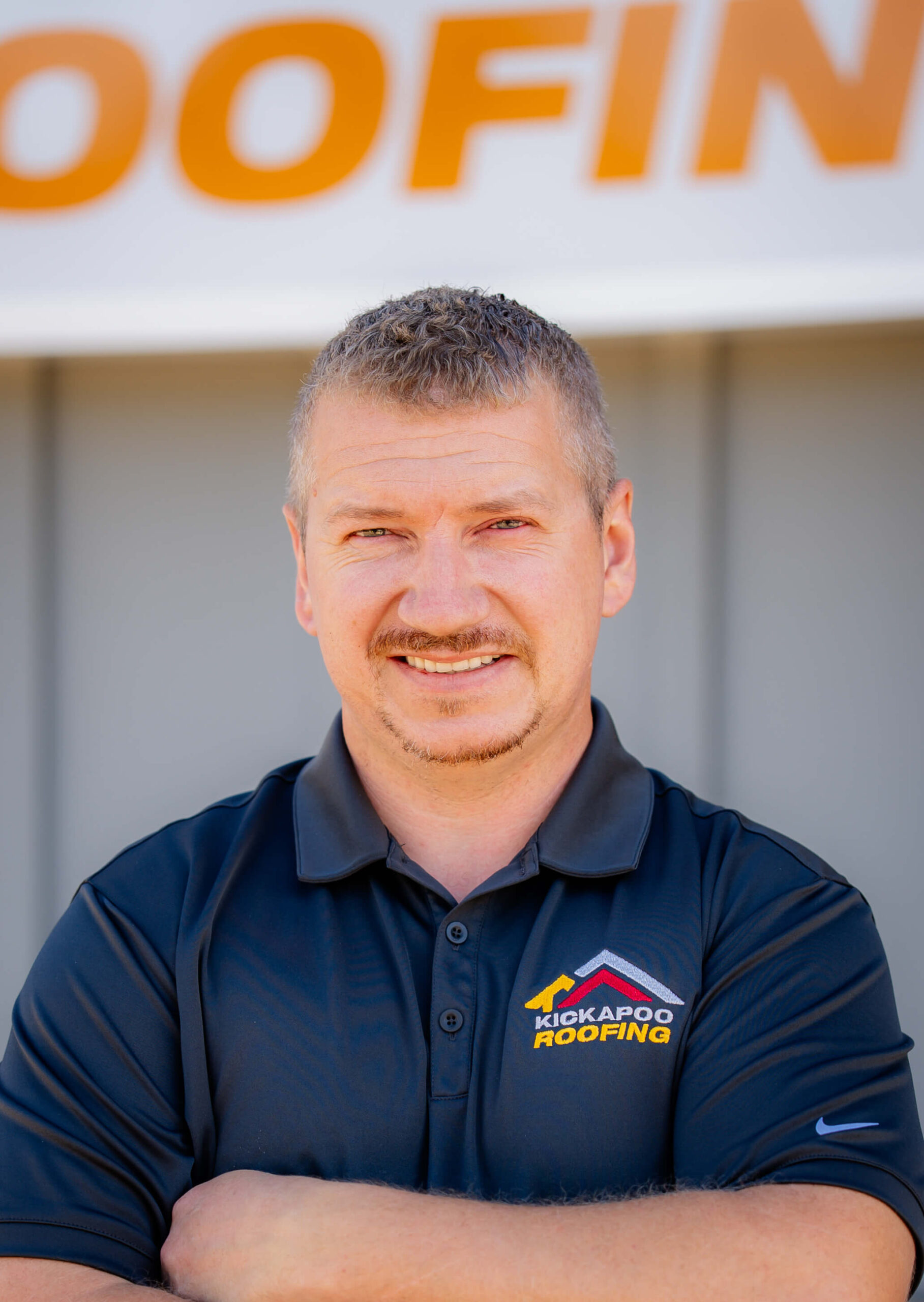 Smiling man in navy Kickapoo Roofing polo shirt with arms crossed outdoors