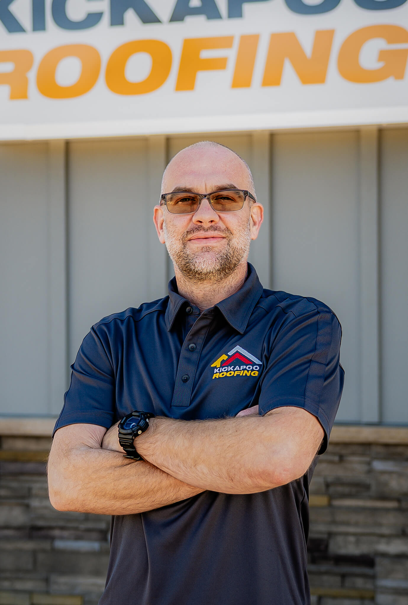 Professional roofing contractor in navy polo shirt with crossed arms outside Kickapoo Roofing building